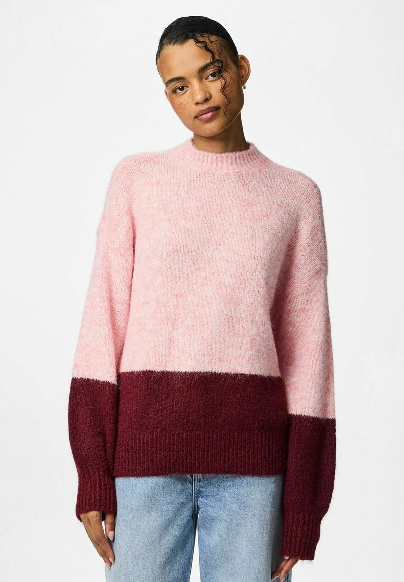 Roza i burgundska prugasta majica s visokim izrezom, dugim rukavima i rebrastim manžetama. Meka, pahuljasta tekstura. Uparen s svijetlop Plavim trapericama.