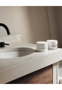 Vasi cilindrici bianchi di crema per il corpo etichettati "Objets d'Amsterdam" su un piano in marmo beige chiaro con un lavabo circolare e un rubinetto nero.