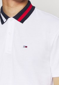 Bílá polo košile s kontrastním námořnickým límcem, který má červené a bílé pruhy, a malým vyšitým logem na levé části hrudi.