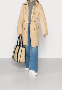 Abrigo impermeable beige, de doble botonadura y con bolsillos laterales. Suéter de cuello alto gris claro, vaqueros de pernera ancha azules y bolso de mano texturizado en negro y beige.