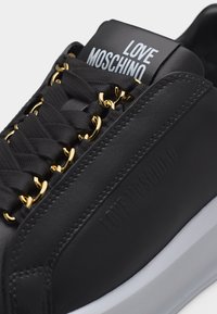 Μαύρο αθλητικό παπούτσι Moschino με χρυσά μεταλλικά στοιχεία, μαύρα κορδόνια και λευκή σόλα, με λεπτομερείς ραφές και λογότυπο στη γλώσσα και το πλάι.