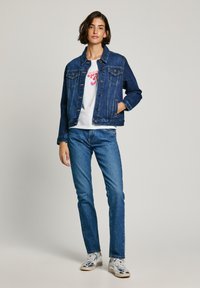 Giacca di denim blu scuro con design classico, due tasche sul petto e orlo grezzo. Abbinata a una maglietta bianca e jeans blu. Sneakers argentate.