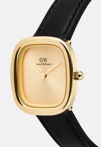 Montre carrée dorée avec un cadran doré brossé et un bracelet en cuir noir. Présente un design minimaliste avec deux aiguilles fines et un détail de couronne.