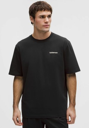 Joven con camiseta negra de manga corta con el logo de "lululemon" en el pecho izquierdo, mirando hacia su izquierda sobre un fondo liso.