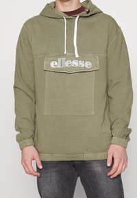 Sudadera de color verde oliva con cordones blancos, bolsillo tipo canguro y logotipo bordado de "ellesse" en el pecho. Presenta puños y dobladillo elásticos.