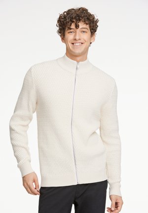 Suéter blanco de punto con cremallera y cuello alto, patrón de diamante texturizado, puños y dobladillo de canalé, llevado por un modelo masculino sonriente.