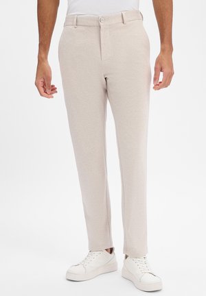 Mann trägt beige, schmal geschnittene Hose mit Knopf und Reißverschluss, kombiniert mit weißen Sneakers und einem weißem, in die Hose gesteckten Hemd.