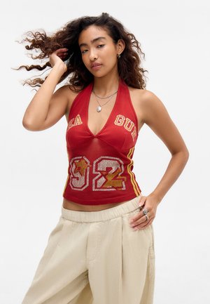 Mladá žena s kučeravými vlasmi, ktorá nosí červený halter crop top s číslom „92“ a béžové široké nohavice, pózuje s rukou vo vlasoch na jednofarebnom pozadí.