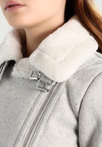 Veste gris clair avec un col blanc en peluche et des accessoires en métal argenté, dotée d'un détail zippé. La texture semble douce et confortable.