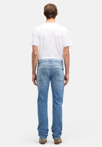 Mężczyzna stojący tyłem, ubrany w prostą białą koszulkę, jasnoniebieskie dżinsy oraz brązowe buty, na tle jednolitej białej ściany.