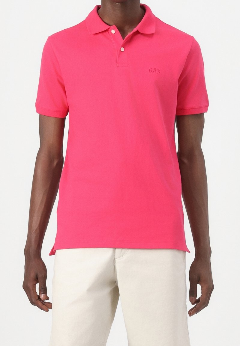 Homme portant un polo rose vif à manches courtes avec deux boutons, associé à un pantalon écru, visible des épaules aux cuisses.
