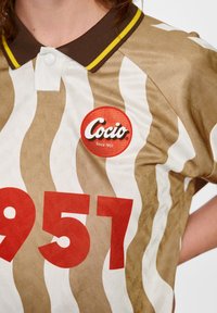 Brun och vit randig skjorta med en brun krage, som har en rund röd "Cocio"-logotyp och siffran "957" i fet röd.