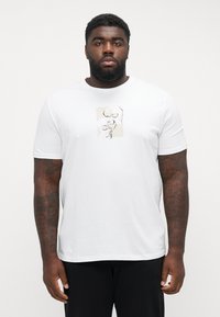 Hvit t-skjorte med kort erm som har et beige og svart abstrakt grafisk motiv foran. Glatt stoff med løs passform og rund hals.