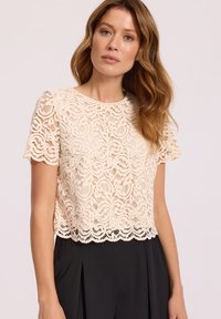 Koronkowy top typu crop w jasnym beżu, z falistym wzorem i falbaniastym dołem. Krótkie rękawy i okrągły dekolt dopełniają całość.