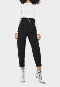 Schwarze hochgeschnittene Hosen mit Falten, mit einem silbernen Schnallen-Gürtel kombiniert, dazu ein weißes Langarmoberteil mit mehrfarbigen Punkten und silberne Knöchelschäfte.