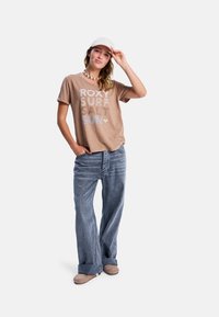 Vrouw in een beige graphic tee, met wijd uitlopende, omgeslagen jeans, beige schoenen, een witte pet en een schelpenhalsketting, staande met één hand op de pet en de andere in haar zak.