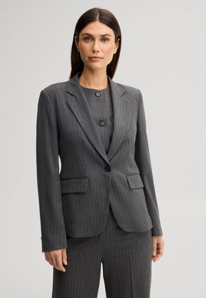 Vrouw met steil bruin haar die een grijs krijtstreepkostuumjasje en bijpassende broek draagt, staand tegen een effen lichte achtergrond.