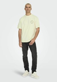 Helles, gelbes Baumwoll-T-Shirt mit Rundhalsausschnitt und kurzen Ärmeln, ausgestattet mit einem kreisförmigen Logo in Marineblau. Kombiniert mit dunklen Jeans und weißen Sneakern.
