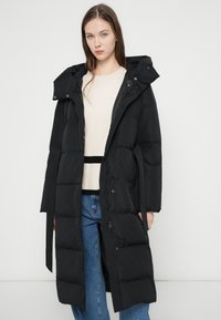 Mladá žena v dlouhém černém péřovém kabátě s kapucí, pod nímž má světlý svetr a modré džíny, stojící před jednobarevným bílým pozadím.