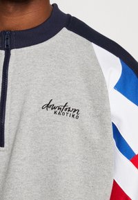 Felpa grigia con collo a zip, con dettagli blu navy e strisce multicolori in rosso, bianco e blu. "downtown KAOTIKO" ricamato sul petto.