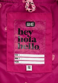 Roza tkanina z teksturirano oznako, ki vsebuje besedilo "hej hola hello", polja za ime in oznako velikosti 122/128. Nastavljiva vrvica na vrhu.