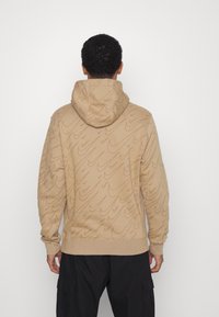 Beige hoodie med en texturerad vågmönster, dragsko i huvan och ribbade ärmmuddar. Tillverkad av mjukt tyg, designad för avslappnad användning.