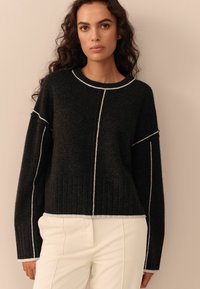 Maglione nero in lana con dettagli bianchi a punto cucito, spalle abbassate e texture a costine all'orlo. Abbinato a pantaloni bianchi in velluto a coste.