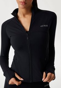 Black zip-up athletic jacket featuring long sleeves, a high collar, and a fitted design. Embroidered "GUESS" logo on the left chest.