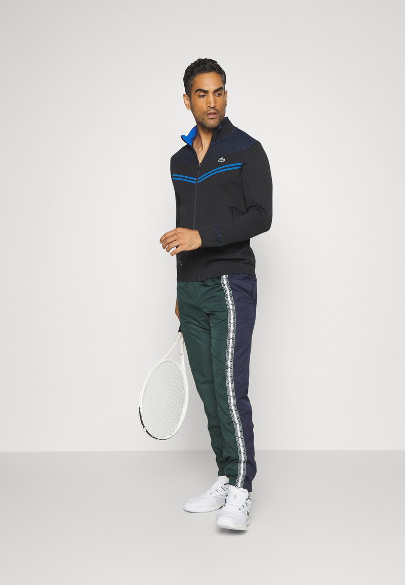 Veste zippée noire avec des accents bleus, associée à un pantalon vert et marine avec un design rayé blanc. Le modèle tient une raquette de tennis.