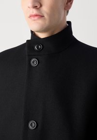 Caban negru din lână cu un guler înalt, prevăzut cu trei nasturi negri, textură netedă, design minimalist și fără modele sau accente vizibile.