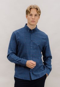 Blå rutete skjorte med knappelukking og krage, laget av et teksturert stoff, med lange ermer og lett buet kant nederst.