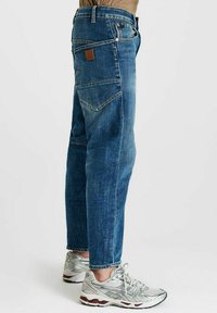 Persoon in relaxed fit blauwe spijkerbroek en zilveren sportschoenen die op een effen witte achtergrond staat.