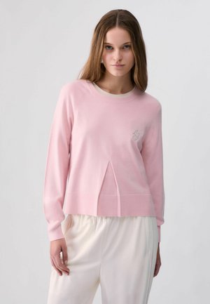 Mujer joven con cabello largo castaño que lleva un suéter rosa claro y pantalones blancos contra un fondo liso y claro.
