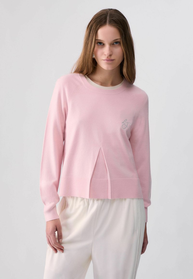 Jeune femme aux longs cheveux bruns portant un pull rose clair et un pantalon blanc sur un fond clair uni.