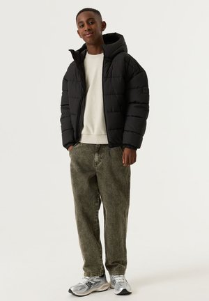 Jonge man staat glimlachend, draagt een zwarte gewatteerde jas, een witte trui, losse grijze jeans en grijze sneakers tegen een egale lichte achtergrond.