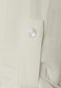 Tessuto color crema con una superficie leggermente strutturata, caratterizzato da una chiusura con bottone cucito sul polsino e un dettaglio con cucitura piegata.