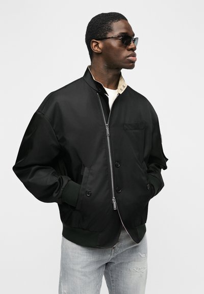 Homme portant une veste bomber noire zippée, jean déchiré bleu clair, lunettes de soleil noires, regardant vers la droite les mains dans les poches.