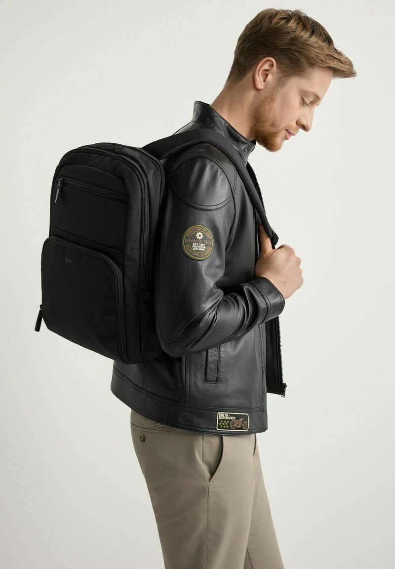 Młody mężczyzna w czarnej skórzanej kurtce i beżowych spodniach niesie czarny plecak na ramieniu, patrząc w dół na tle gładkiego tła.