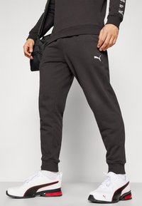 Schwarze Sweatpants mit schmalem Schnitt, aus weichem Stoff, mit einem weißen Puma-Logo. Kombiniert mit weißen Schuhen mit schwarzen und roten Akzenten.