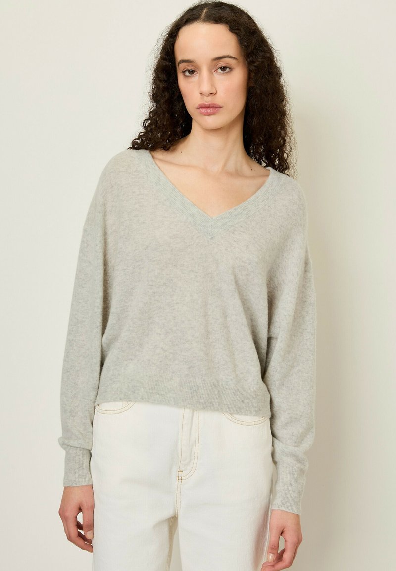 Pull en v léger gris fabriqué à partir d'un tissu doux et texturé, avec des manches longues et une coupe légèrement cropped, associé à un jean blanc.