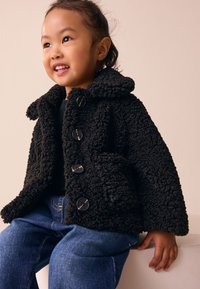 Black fleece jacket with a textured surface, large buttons, and a hood. Paired with blue denim jeans, showcasing a relaxed fit.