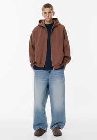 Hnedá bunda na zips s veľkou kapucňou cez námornícku košeľu, doplnená širokými svetlomodrými džínami a sivými nazúvacími topánkami.