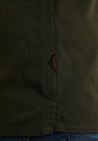 Tissu texturé vert foncé avec une couture verticale et une petite étiquette noire et orange, porté par-dessus un jean en denim bleu.