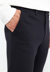 Zwarte broek met een gladde textuur, zijzak en ritsluiting. De tailleband is standaard en de stof lijkt duurzaam te zijn.