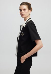 Czarna krótka kurtka z krótkim rękawem i białym kołnierzykiem oraz zamkiem błyskawicznym, z haftowanym złotym logo i minimalistycznym wzornictwem.
