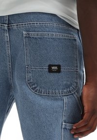 Pantaloni cargo in denim azzurro chiaro con numerose tasche, cuciture in evidenza e un patch del logo Vans nero sulla tasca posteriore.