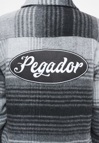 Camicia in flanella a quadri grigia con un grande pannello ovale nero sul retro, con il testo bianco "Pegador". Tessuto morbido con diverse sfumature.