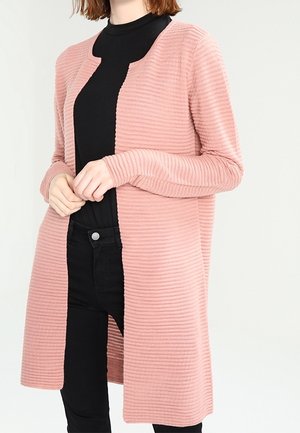 Vrouw draagt een lange, geribde roze cardigan over een zwart shirt en zwarte broek, houdt de randen van de cardigan met beide handen vast.