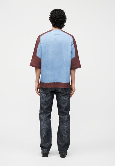 Άνδρας με σγουρά μαλλιά, φορώντας ένα φαρδύ μπλε και καφέ κοντομάνικο πουκάμισο και σκούρο παντελόνι με σχέδια, στέκεται με την πλάτη προς τα εμπρός σε λευκό φόντο.