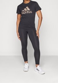 Svart bomullst-shirt med Adidas-logga i leopardmönster, kombinerad med svarta leggings och vita sneakers. Outfiten har en figurnära design.